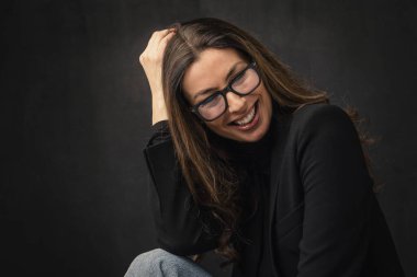 Headshot of an attractive middle aged woman with toothy smile wearing turtleneck sweater while sitting at isolated dark background. Copy space. Studio shot. Hand on forehead. 