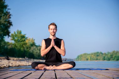 Göl kenarındaki iskelede yoga yapan beyaz bir adam. Olgun bir adam, mavi gökyüzüne karşı iskelede nilüfer pozisyonu alıyor..