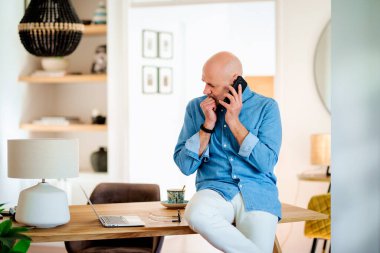 Confident middle aged man making a call and using laptop while working from home. Confident male sitting in the dining room and having a call. Home office.