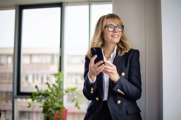 A middle-aged businesswoman standing in the office and using smartphone. Confident professional woman wearing blazer and text messaging.