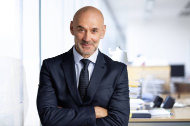 Ofiste duran orta yaşlı iş adamı. Takım elbiseli, kravatlı ve kameraya bakan kendine güvenen bir adam.. 