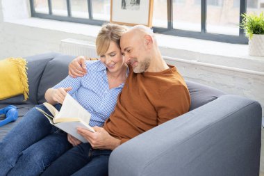 Evli bir çiftin birlikte kitap okuduğu ve evdeki kanepede dinlendiği bir fotoğraf. Sarışın kadın ve günlük kıyafetler giyen orta yaşlı adam.. 