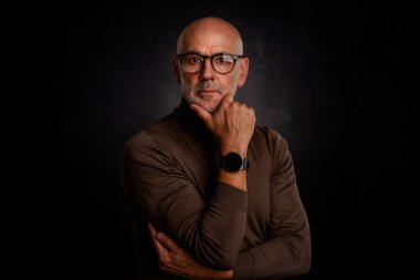 Gözlüklü olgun bir adamın portresi izole edilmiş siyah arka plana karşı düşünceli görünüyor. Balıkçı yaka kazak giyen kendinden emin bir erkek..