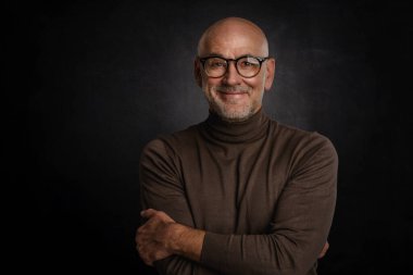 Siyah arka planda kameraya gülümseyen gözlüklü olgun bir adamın portresi. Balıkçı yaka kazak giyen kendinden emin bir erkek..