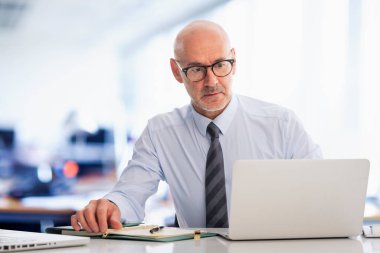 Ciddi yüzlü iş adamı ofiste oturuyor ve not defterini iş için kullanıyor. Orta yaşlı profesyonel bir adam düşünceli görünüyor.. 