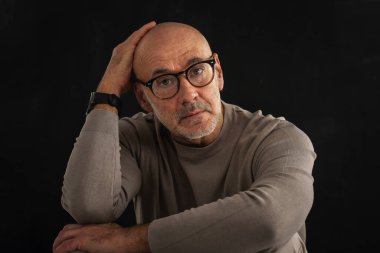 Orta yaşlı bir adam düşünceli görünüyor ve siyah arka planda oturuyor. Kel ve sakallı bir adam, izole bir arka planda günlük kıyafetler ve gözlük takıyor. Boşluğu kopyala. 