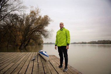 İskelede duran ve kanoya binmeye hazır orta yaşlı bir adam. Aktif sporcu kuru takım elbise giyiyor ve kameraya bakıyor.. 