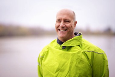 Orta yaşlı, kuru takım elbiseli ve nehir kıyısındaki iskelede duran bir adamın yakın plan portresi. Başka tarafa bakıyor ve gülüyor.