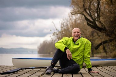 İskelede oturan ve kanoya binmeye hazır orta yaşlı bir adam. Aktif sporcu kuru takım elbise giyiyor ve kameraya bakıyor.. 
