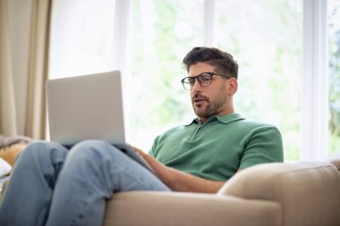 Orta yaşlı bir adam evdeki koltuğunda oturup iş için dizüstü bilgisayar kullanıyor ya da video görüşmesi yapıyor. Gündelik kıyafetler giyen kendine güvenen bir iş adamı. Merkez Ofis.