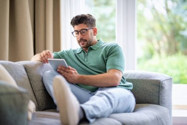 Orta yaşlı bir adam evde oturup dijital tablet kullanıyor. İnternette geziniyor ya da video bağlantısı yok..