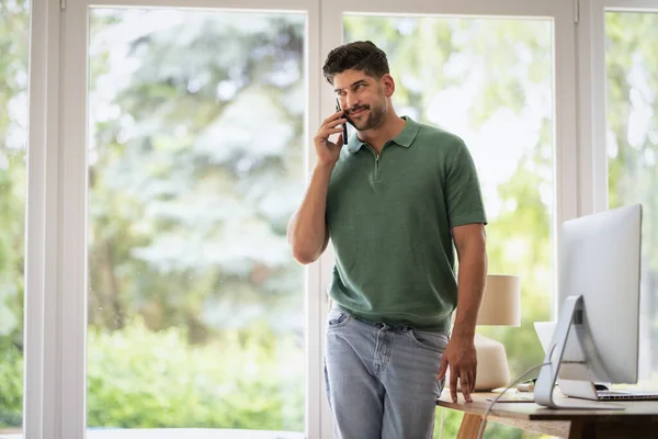 A confident man standing at the desk in the office and making a phone call. Professional male wearing casual clothes and working from home. Home office.