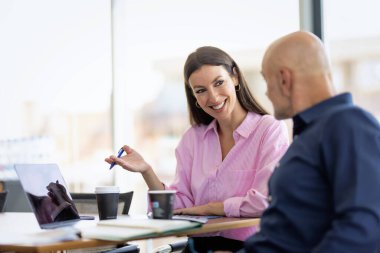 Bir grup iş kadını ve bir iş adamı modern bir ofiste iş toplantısındalar. Masada dizüstü bilgisayar ve belgeler var..