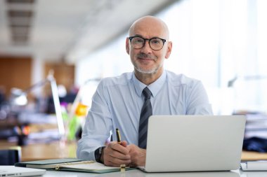 Ofiste oturan orta yaşlı bir işadamı ve not defterini iş için kullanıyor. Gömlek ve kravat takan kendinden emin profesyonel bir adam.. 