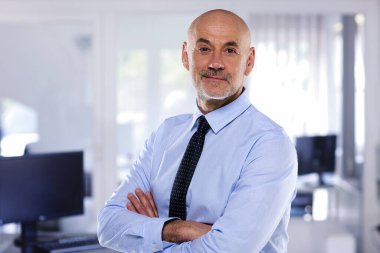 Ofiste duran kendinden emin orta yaşlı işadamı. Profesyonel erkek gömlek ve kravat takıyor ve kolları çapraz duruyor.. 