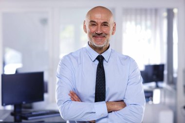 Ofiste duran kendinden emin orta yaşlı işadamı. Profesyonel erkek gömlek ve kravat takıyor ve kolları çapraz duruyor.. 