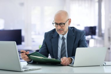 Ofisindeki masasında oturmuş dizüstü bilgisayar kullanan orta yaşlı bir iş adamı. Elinde not defteri tutan bir yönetici ve düşünceli görünüyor..