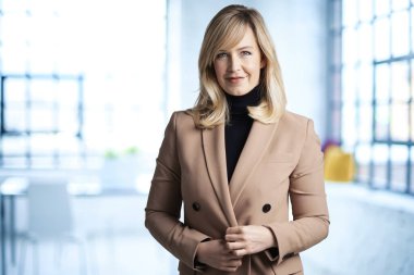 Ofiste duran orta yaşlı, sarı saçlı bir kadın. Bir ceket giyiyor ve kameraya gülümsüyor.. 