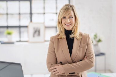 Ofiste duran orta yaşlı, sarı saçlı bir kadın. Bir ceket giyiyor ve kameraya gülümsüyor.. 