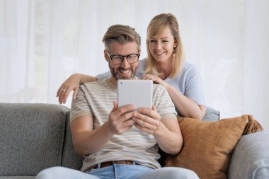 Orta yaşlı bir kadın ve erkek kanepede oturuyorlar ve dijital tablet kullanıyorlar. Sarışın, kadın ve yakışıklı adam günlük kıyafetler giyiyor ve gülümsüyor.. 