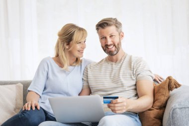 Evden online alışveriş. Karısı yanında oturmuş internetten alışveriş yaparken elinde banka kartı tutan yakışıklı bir adam.. 