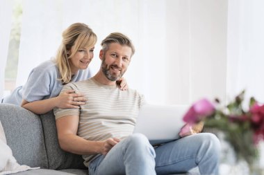 Orta yaşlı bir kadın ve erkek evde kanepede oturuyorlar ve dizüstü bilgisayar kullanıyorlar. Sarışın, kadın ve yakışıklı adam günlük kıyafetler giyiyor ve gülümsüyor.. 