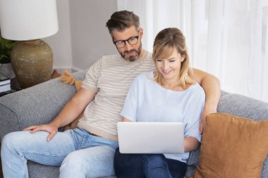 Orta yaşlı bir kadın ve erkek evde kanepede oturuyorlar ve dizüstü bilgisayar kullanıyorlar. Sarışın, kadın ve yakışıklı adam günlük kıyafetler giyiyor ve gülümsüyor.. 