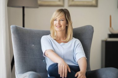 Sarışın, çekici bir kadın. Evdeki koltukta dinleniyor. Orta yaşlı, neşeli, gülümseyen ve hayal kuran bir kadın..