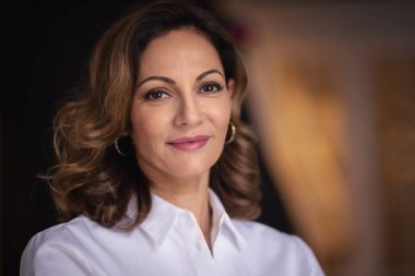 Sarışın, orta yaşlı bir kadının kapalı mekanda otururken yakın plan portresi. Gri kazak giyen ve kameraya gülümseyen kendine güvenen bir kadın.. 