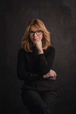 Sarışın, orta yaşlı, karanlık arka planda stüdyo resmi olan bir kadın. Siyah kazak ve siyah pantolon giyen kendinden emin bir kadın.. 