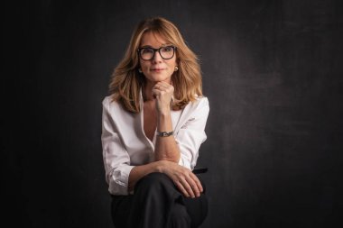 Sarışın, orta yaşlı, karanlık arka planda stüdyo resmi olan bir kadın. Beyaz gömlek ve siyah pantolon giyen kendinden emin bir kadın.. 