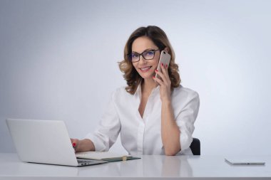 Orta yaşlı bir iş kadını, izole edilmiş beyaz arka planda masa başında oturuyor. İş için dizüstü bilgisayar ve cep telefonu kullanan kendine güvenen profesyonel bir kadın. Boşluğu kopyala.