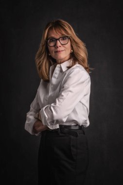 Sarışın, orta yaşlı, karanlık arka planda stüdyo resmi olan bir kadın. Beyaz gömlek ve siyah pantolon giyen kendinden emin bir kadın.. 
