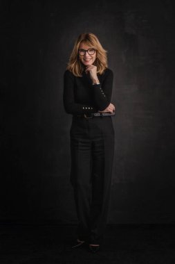 Sarışın, orta yaşlı, karanlık arka planda stüdyo resmi olan bir kadın. Siyah kazak ve pantolon giyen kendinden emin bir kadın. Tam boy çekim.