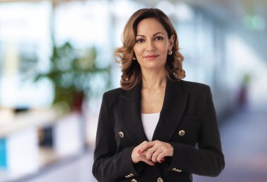 Ofis koridorunda duran sarı saçlı bir iş kadını. Orta yaşlı profesyonel kadın siyah ceket giyiyor ve kameraya bakıyor. Boşluğu kopyala.