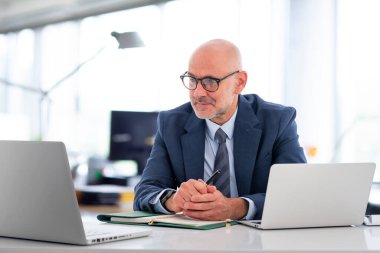 Ofiste oturan orta yaşlı bir işadamı ve not defterini iş için kullanıyor. Kendine güvenen profesyonel adam video konferansı yapıyor.. 