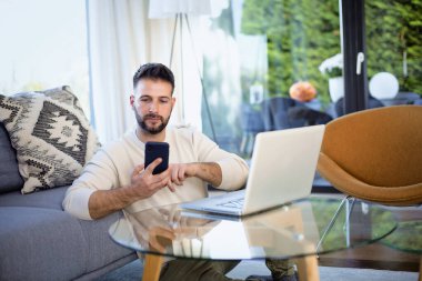 Evde dizüstü bilgisayar ve cep telefonu kullanan orta yaşlı bir adam. Kendine güveni tam bir erkek, günlük kıyafetler giyiyor ve evden çalışıyor. Merkez Ofis.