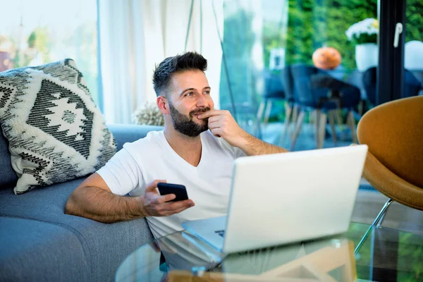 A mid aged man sitting at home using a laptop and mobile phone. Confident male wearing casual clothes and working from home. Home office.