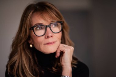 Sarışın, orta yaşlı, evin içinde dikilen kadının yakın plan portresi. Siyah kazak giyen ve düşünceli görünen kendine güvenen bir kadın..