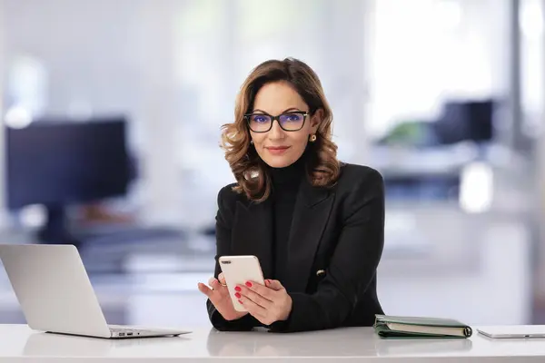 Mid age businesswoman sitting at the office and using laptop and smartphone for work. Confident professional female wearing black jacket. Copy space.