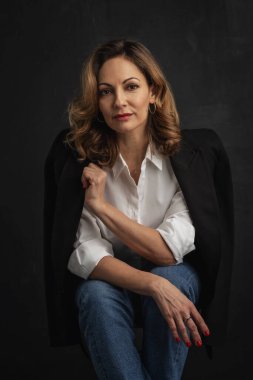 Koyu renk arka planda sarı saçlı bir kadın stüdyo resmi. Beyaz gömlek ve kot pantolon giyen orta yaşlı bir kadın..