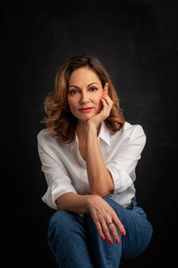 Sarışın, orta yaşlı, karanlık arka planda stüdyo resmi olan bir kadın. Beyaz gömlek ve kot pantolon giyen kendinden emin bir kadın..