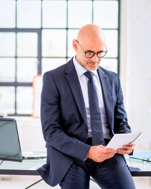 Orta yaşlı bir işadamı ofisinde oturuyor. Takım elbiseli ve kravatlı kendine güvenen profesyonel bir adam..