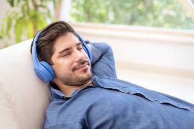 Kulaklığı elinde, evdeki kanepede uzanıp müzik dinleyen bir adamın yakın çekimi..