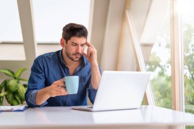 Bir adam masada oturuyor ve dizüstü bilgisayar kullanıyor. O, düşünceli görünüyor. Kendine güveni tam bir erkek, günlük kıyafetler giyiyor ve çay içiyor. Merkez Ofis. 
