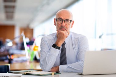 Ofiste oturan orta yaşlı bir iş adamı. Adamın ciddi bir yüzü var ve tişört giyiyor..