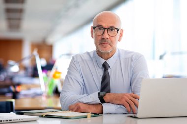 Ofiste oturan orta yaşlı bir iş adamı. Adamın ciddi bir yüzü var ve tişört giyiyor..