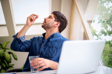 İlaç alan bir adam. Orta yaşlı bir adam evde dizüstü bilgisayarının önünde oturup ilaçlarını alıyor. Günlük giysiler giyiyor ve evden çalışıyor. Merkez Ofis.