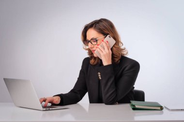 Orta yaşlı bir iş kadını, izole edilmiş beyaz arka planda masa başında oturuyor. İş için dizüstü bilgisayar ve cep telefonu kullanan kendine güvenen profesyonel bir kadın. Boşluğu kopyala.