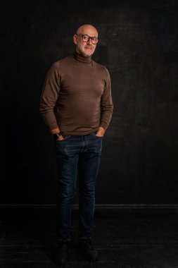 Tecrit edilmiş karanlık arka plana karşı duran yaşlı bir adamın boyu. Balıkçı yaka kazak ve kot pantolon giyen kendinden emin bir erkek. Uzayı kopyala Ciddi yüz.. 